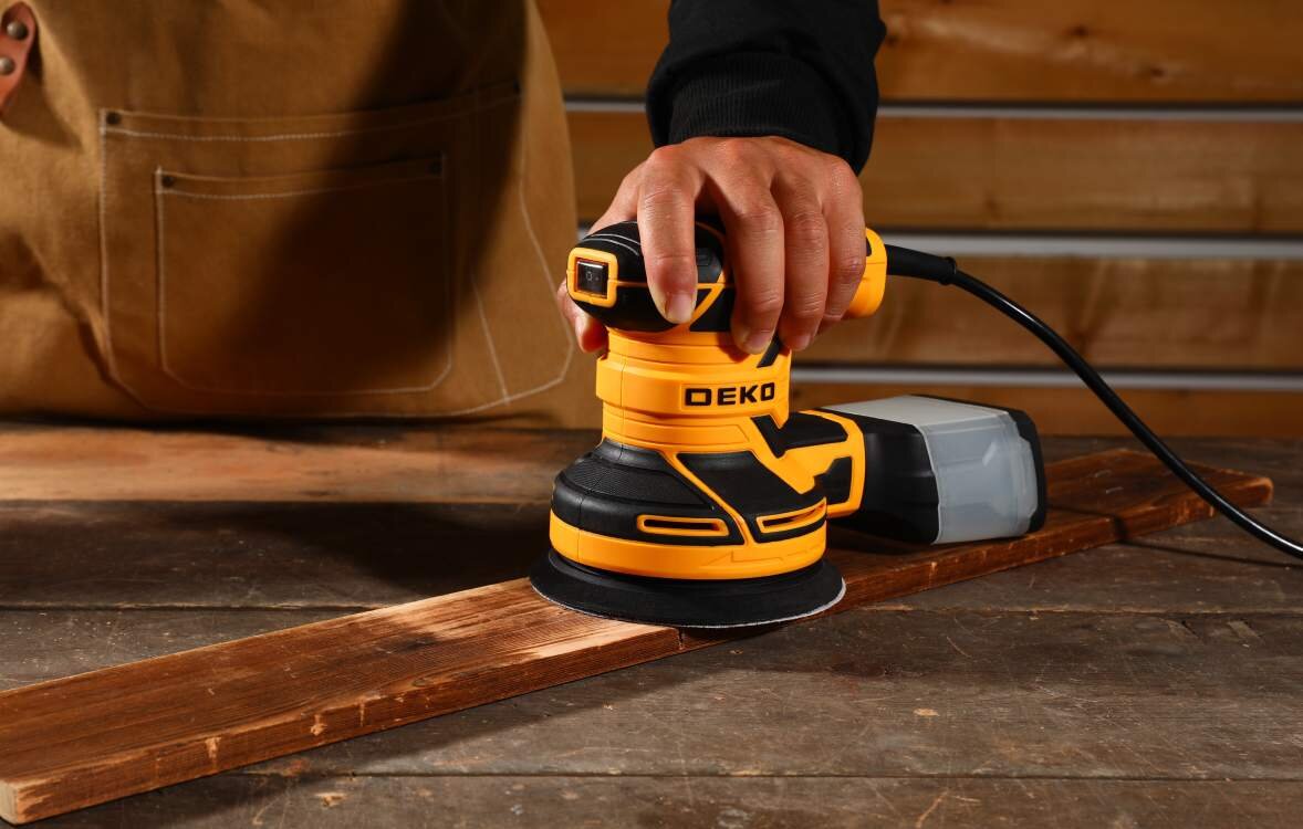 Osoba używa szlifierki mimośrodowej DEKO na drewnianej desce. Szlifierka jest żółto-czarna z widocznym logo \'DEKO\'.
