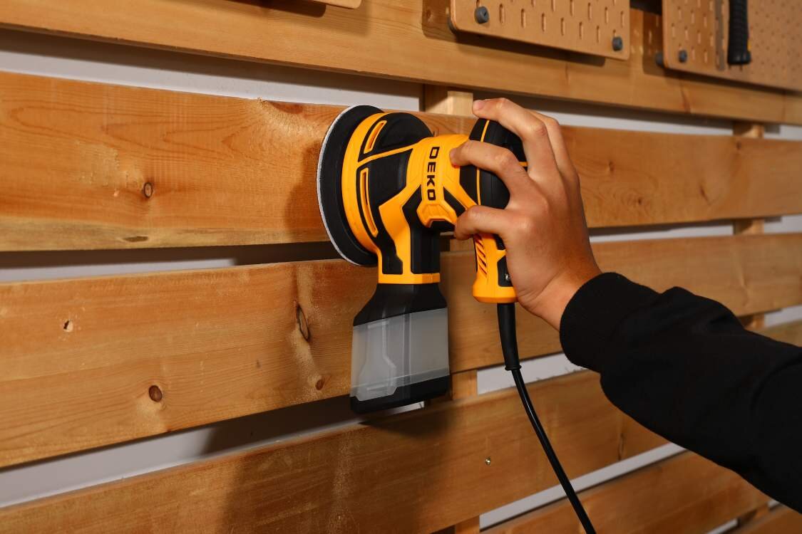 Osoba używa szlifierki mimośrodowej DEKO na drewnianej powierzchni. Urządzenie jest w kolorze żółto-czarnym z widocznym logo \'DEKO\'.