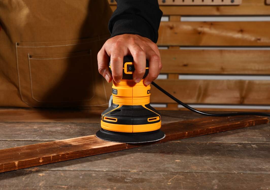 Osoba używa żółto-czarnej szlifierki mimośrodowej na drewnianej desce. W tle widoczna jest drewniana ściana z narzędziami.