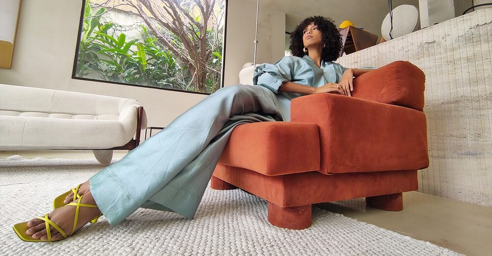 Osoba w jasnoniebieskim stroju siedzi na pomarańczowym fotelu w nowoczesnym wnętrzu z dużym oknem i roślinnością na zewnątrz. Na podłodze leży jasny dywan, a w tle widać biały fotel.