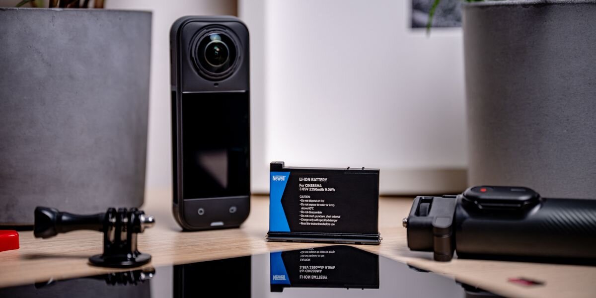 Akumulator NEWELL 2350 mAh Cinsbbma do Insta360 X4, Czarna bateria Newell stoi pionowo obok kamery 360 i uchwytu na blacie. W tle rośliny w szarych doniczkach, 2350mAh, Li-Ion, 9,9Wh, nagranie