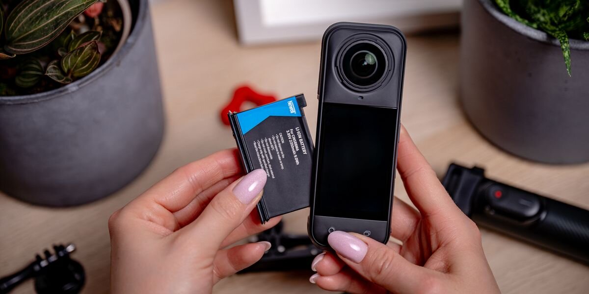 Akumulator NEWELL 2350 mAh Cinsbbma do Insta360 X4, Dłonie trzymają czarną baterię Newell obok kamery 360. W tle akcesoria foto i zielone rośliny w doniczkach, precyzyjny, komora, obudowa, bezpamięciowy