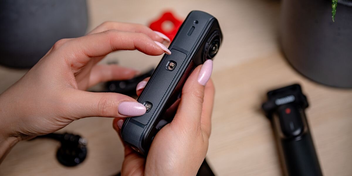 Akumulator NEWELL 2350 mAh Cinsbbma do Insta360 X4, Dłonie wsuwają czarną baterię Newell do bocznego gniazda kamery 360. Widoczna chropowata faktura obudowy urządzenia, przegrzanie, przeciążenie, przeładowanie, zabezpieczenie