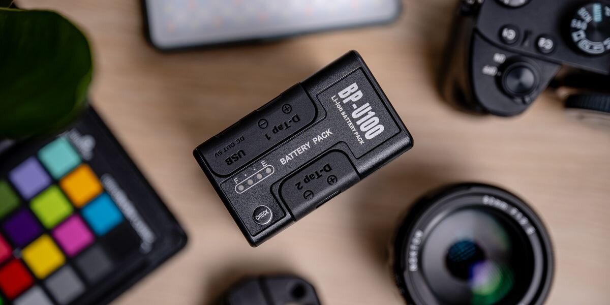 Akumulator NEWELL 6700 mAh BP-U100 do Sony, Czarny akumulator Newell BP-U100 z napisami i dwoma gniazdami D-Tap, leżący na blacie obok aparatu i obiektywu, praca, zasilanie, ładowanie, zastąpienie