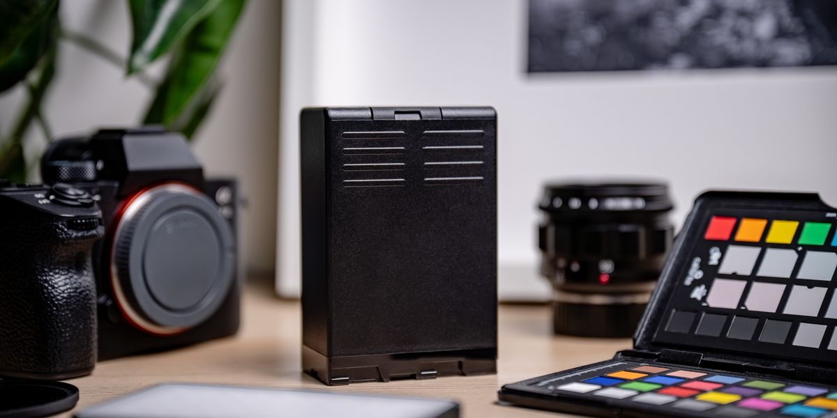 Akumulator NEWELL 6700 mAh BP-U100 do Sony, Pionowe ujęcie boku czarnego akumulatora BP-U100 stojącego na biurku w otoczeniu sprzętu fotograficznego, Li-ion, bezpamięciowy, doładowywanie, całodniowy