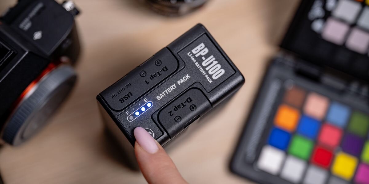 Akumulator NEWELL 6700 mAh BP-U100 do Sony, Dłoń naciskająca przycisk „Check” na akumulatorze, co podświetla cztery niebieskie diody wskaźnika poziomu naładowania, litowo-jonowy, wielokrotny, wydajność, twórczy