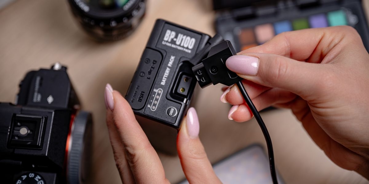 Akumulator NEWELL 6700 mAh BP-U100 do Sony, Dłonie trzymające akumulator BP-U100 i podłączające czarny wtyk do odsłoniętego gniazda D-Tap na jego obudowie, IC, przegrzanie, zwarcię, monitorowanie