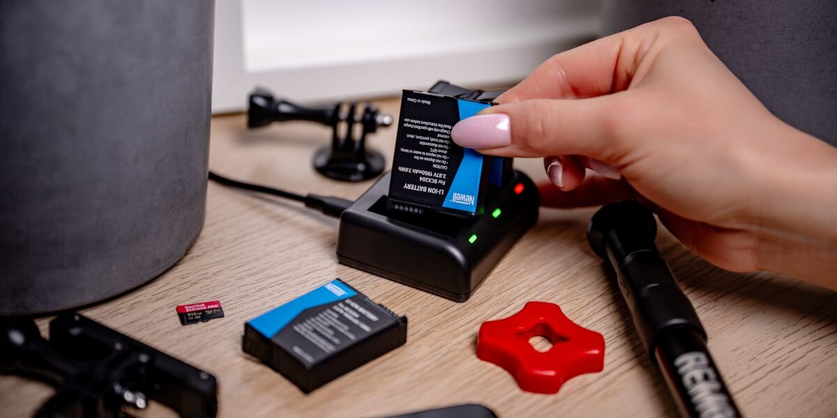 Ładowarka trzykanałowa NEWELL SDC-USB-C + 3 akumulatory BCX-204 do DJI Osmo Action, Dłoń wkłada akumulator do ładowarki, obok widoczne karty pamięci i uchwyty do kamery na biurku, energia, ładowarka, akumulatory, kamera