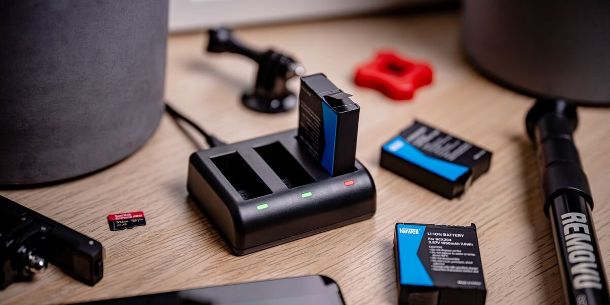 Ładowarka trzykanałowa NEWELL SDC-USB-C + 3 akumulatory BCX-204 do DJI Osmo Action, Ładowarka z jednym akumulatorem, diody LED świecą, wokół leżą akcesoria i karta pamięci, baterie, nagrywanie, zapas, działanie