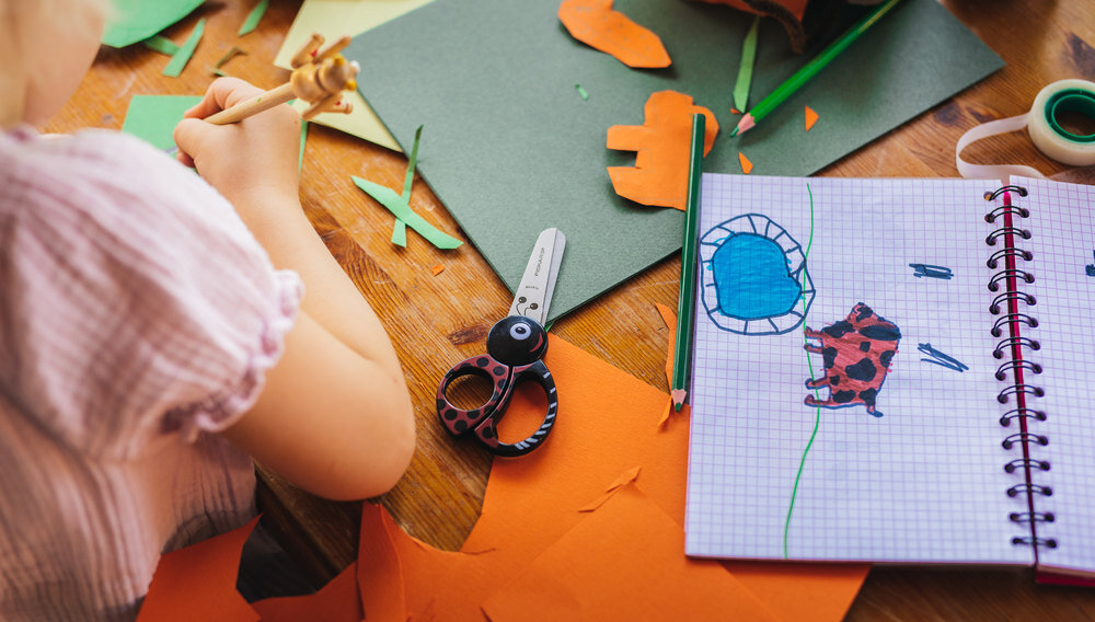 Na stole leżą kolorowe kartki papieru, nożyczki z czarnymi uchwytami w czerwone kropki oraz otwarty zeszyt z rysunkiem biedronki i niebieskiego kształtu. Obok zeszytu znajduje się zielony ołówek.