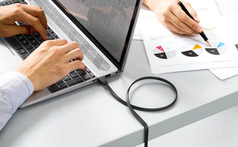 Na biurku znajduje się laptop z podłączonym kablem RJ-45, obok leży dokument z wykresami kołowymi, a osoba trzyma długopis nad kartką.