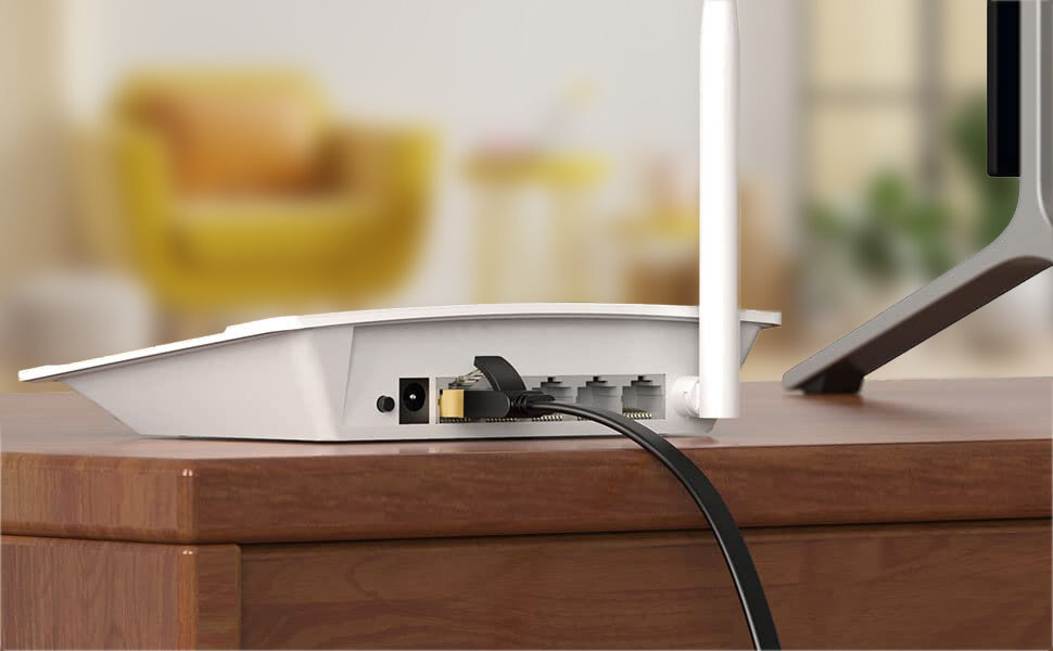 Na drewnianym stole znajduje się biały router z podłączonym czarnym kablem RJ-45. W tle widoczny jest rozmyty obraz wnętrza z żółtym fotelem.