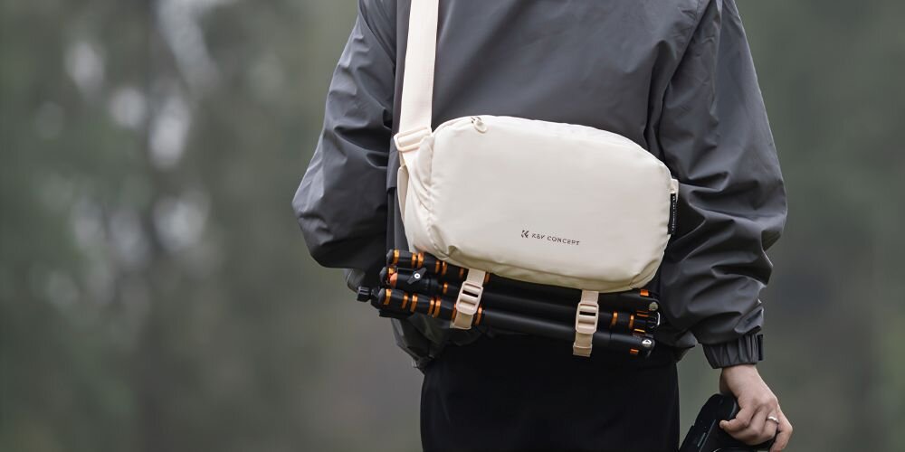 Młody mężczyzna stojący tyłem ma przewieszoną przez tułów torbę Alpha Sling Bag. Na spodzie torby przymocowany jest statyw