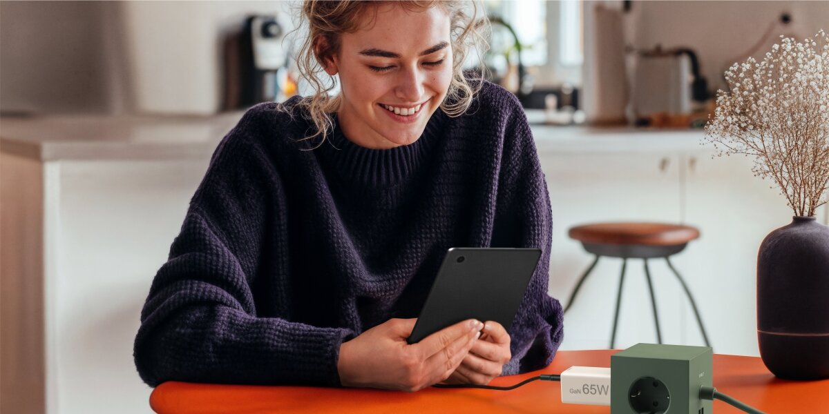 Osoba siedzi przy stole, trzymając tablet. Na stole znajduje się ładowarka z napisem \'65W\'.