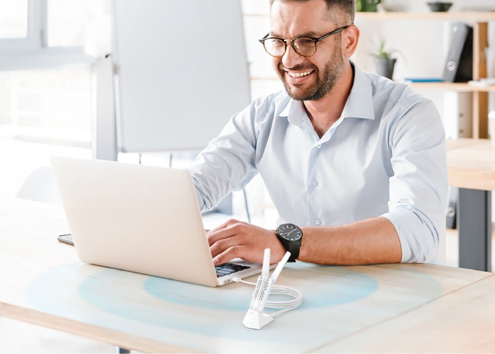 Karta sieciowa D-LINK AE65U Mężczyzna w okularach i jasnej koszuli uśmiecha się pracując przy laptopie na drewnianym biurku, przed którym stoi biała karta sieciowa w podstawce z naniesionym graficznym symbolem rozchodzących się błękitnych fal