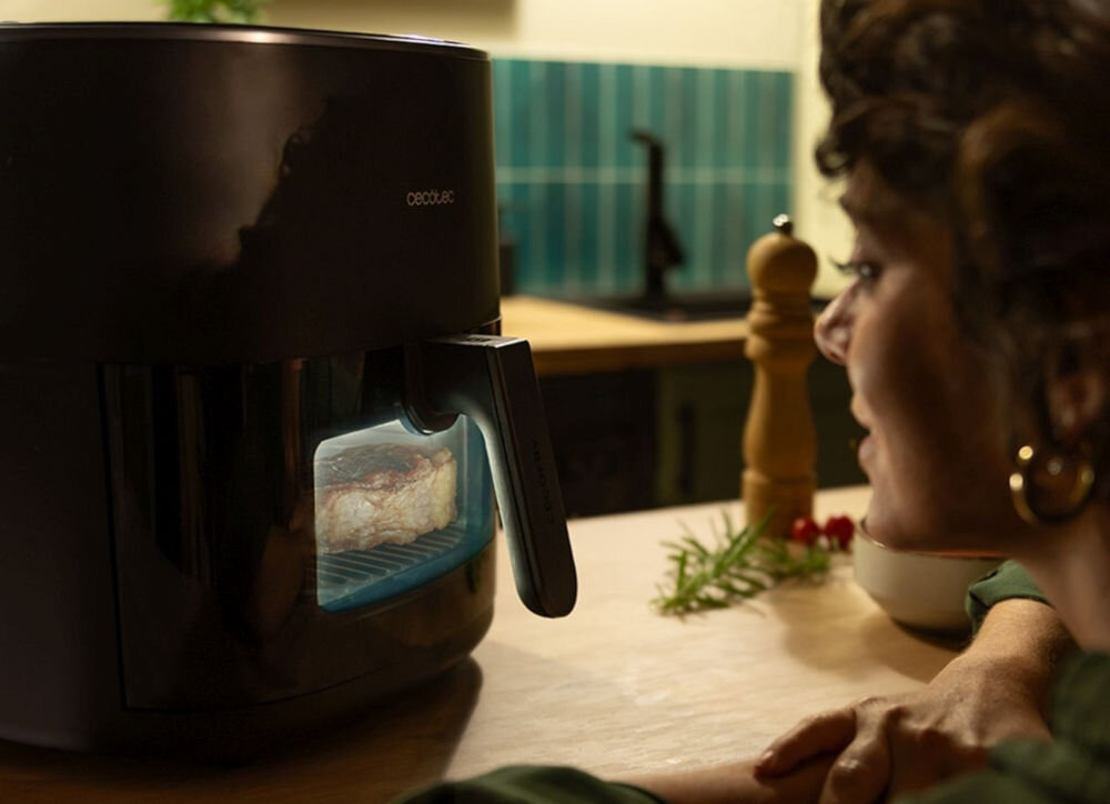 Air Fryer Frytkownica beztłuszczowa CECOTEC CecoFry&Grill Duoheat 6500 3W1 z podwójną grzałką osoba oparta o blat patrzy w świecące okienko w urządzeniu uchwyt Panoramiczne okienko śledzenie procesu gotowania temperatura wewnątrz pozostaje stabilna Możliwość monitorowania jedzenia oszczędność czasu i energii 