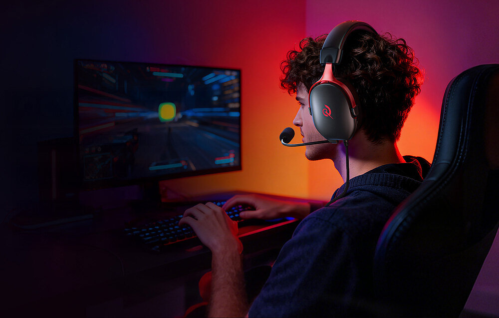 Osoba siedzi przy biurku, grając na komputerze w grze wideo. Na głowie ma czarno-czerwone słuchawki z mikrofonem.