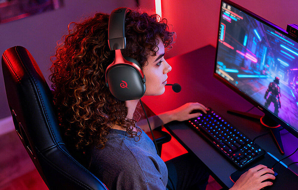 Osoba siedzi przy biurku, grając na komputerze, nosząc czarno-czerwone słuchawki z mikrofonem. Na ekranie widoczna jest gra w futurystycznym stylu z neonowymi kolorami.