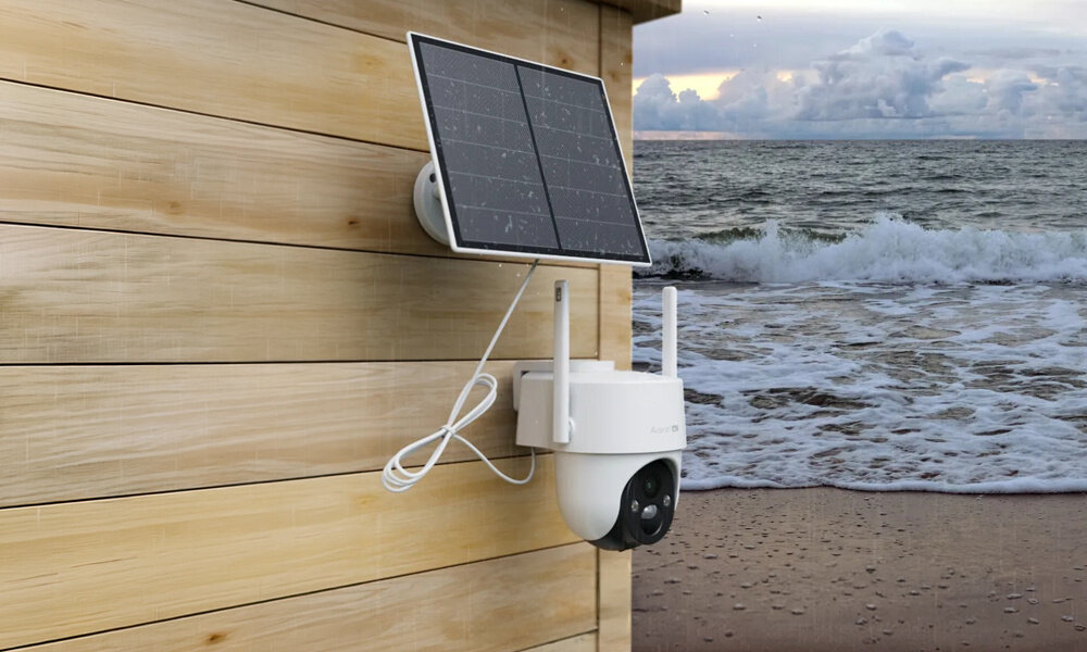 Na drewnianej ścianie zamontowana jest kamera z panelem solarnym. W tle widać wzburzone morze i plażę.