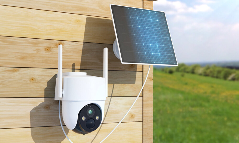 Kamera Arenti z panelem solarnym jest zamontowana na drewnianej ścianie budynku. W tle znajduje widać zieloną polanę.