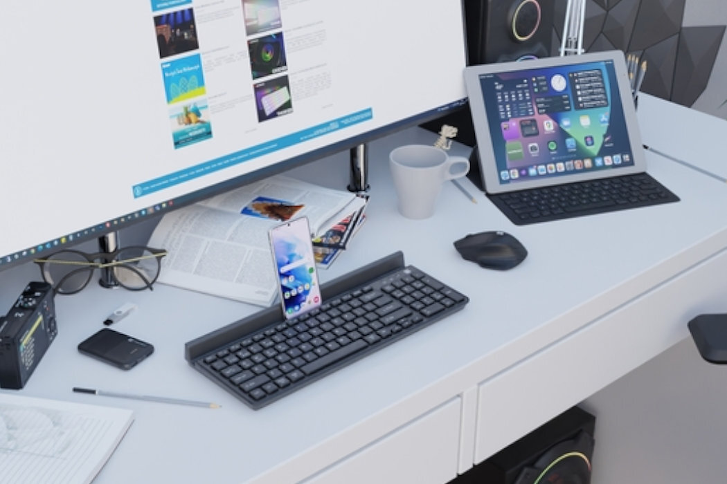 Na biurku znajduje się klawiatura, mysz, smartfon, tablet z klawiaturą oraz monitor wyświetlający stronę internetową. Obok leżą okulary, kubek, długopis i otwarta książka.