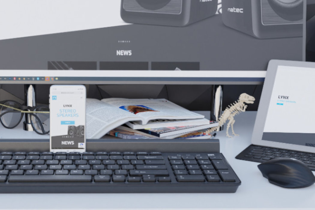 Na biurku znajduje się klawiatura, myszka i telefon z wyświetlonym tekstem \'LYNX STEREO SPEAKERS NEWS\'. W tle widoczny jest monitor z logo \'natec\' oraz otwarta książka i figurka szkieletu dinozaura.