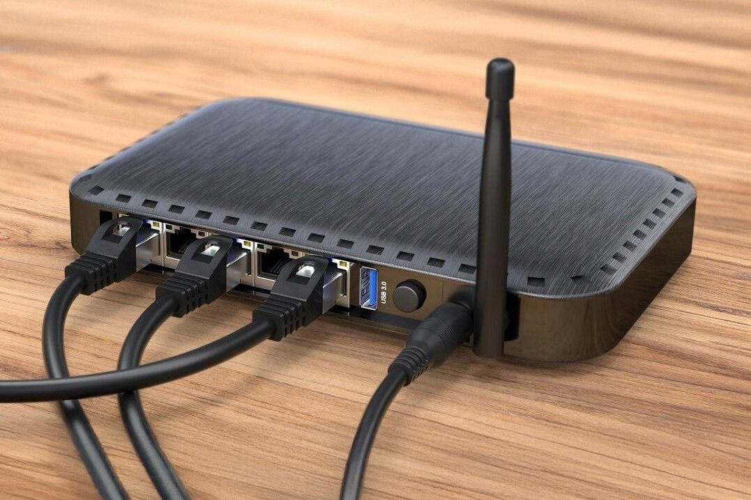 Na drewnianym blacie znajduje się czarny router z kilkoma podłączonymi kablami sieciowymi i anteną. Obok portów widoczny jest napis \'RESET\'.