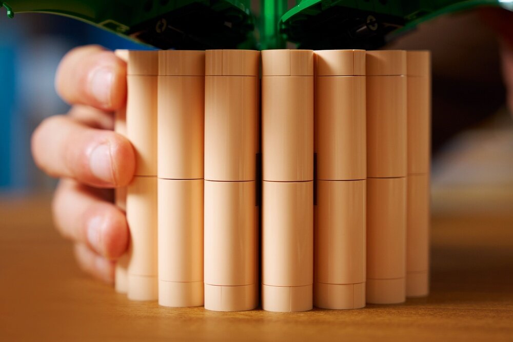 Zdjęcie przedstawia rękę trzymającą cylindryczną konstrukcję z jasnobrązowych elementów LEGO. W tle widoczne są zielone elementy przypominające liście.