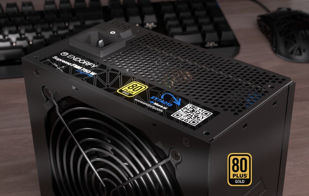 Komputer MAD DOG ENDORFY500ARGB-A35WR32WP Zbliżenie na zasilacz ustawiony na biurku. Widoczna jest naklejka informacyjna z oznaczeniem modelu, certyfikatem 80 PLUS Gold, kodem QR. W tle widać fragment klawiatury i myszy komputerowej, zasilacz, moc, sprawność, kondensatory