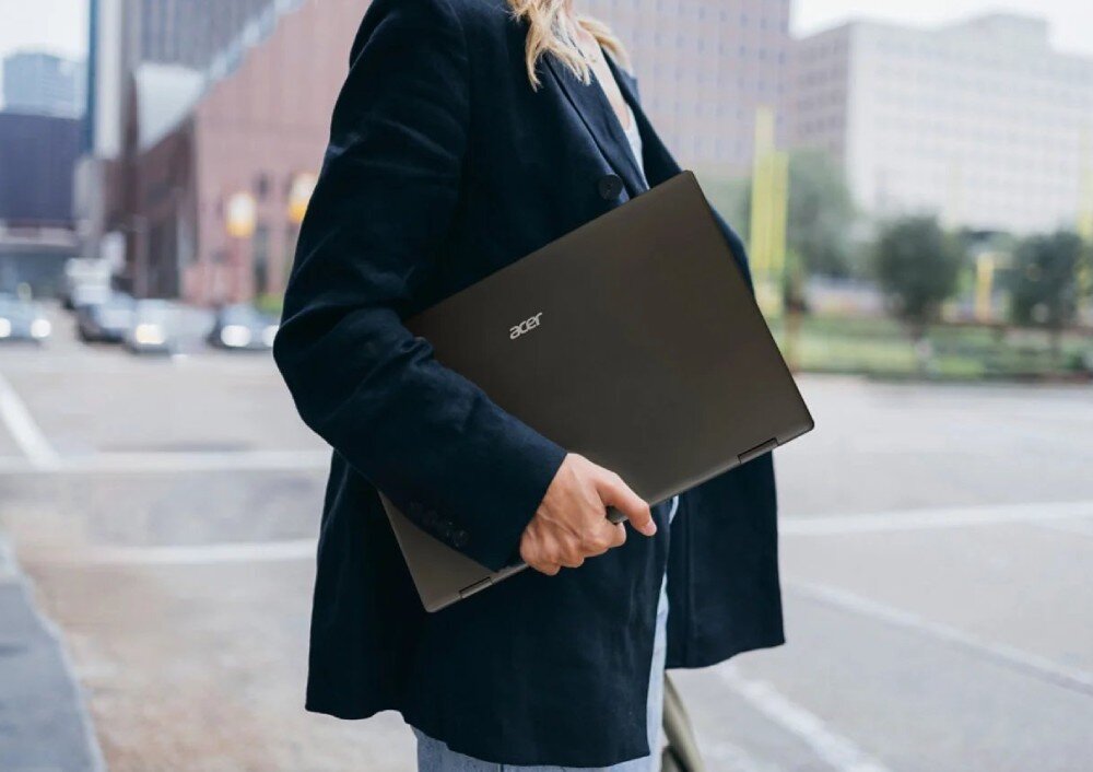 Monitor ACER PD163QT Kobieta w ciemnej marynarce idąca miejską ulicą i niosąca pod pachą złożone, płaskie urządzenie z widocznym małym logo na obudowie