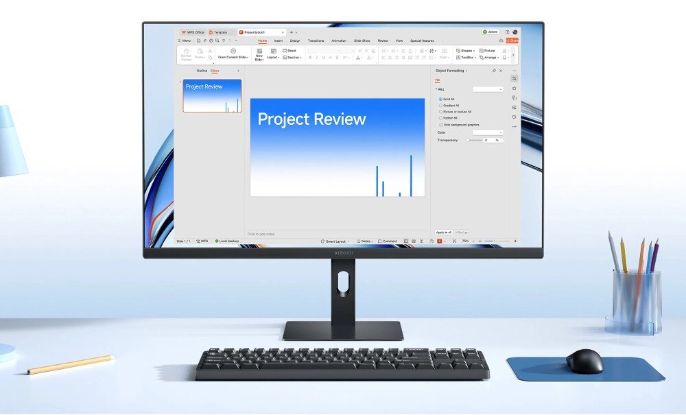 Na biurku znajduje się monitor wyświetlający prezentację z tytułem \'Project Review\'. Przed monitorem leży klawiatura, a obok myszka na podkładce oraz pojemnik z kolorowymi długopisami.