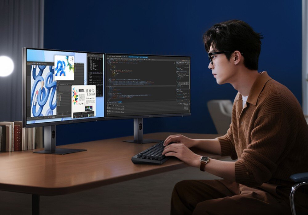 Monitor XIAOMI A27QI 2026 Mężczyzna w okularach siedzi przy drewnianym biurku i pisze na klawiaturze korzystając z dwóch monitorów ustawionych obok siebie, wygląd, konstrukcja, mocowanie VESA