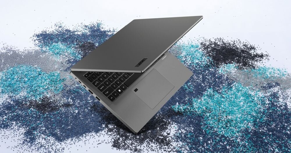 Na obrazie widoczny jest otwarty laptop w kolorze szarym, umieszczony na tle kolorowych drobinek w odcieniach niebieskiego i czarnego. Na obudowie laptopa znajduje się logo producenta.