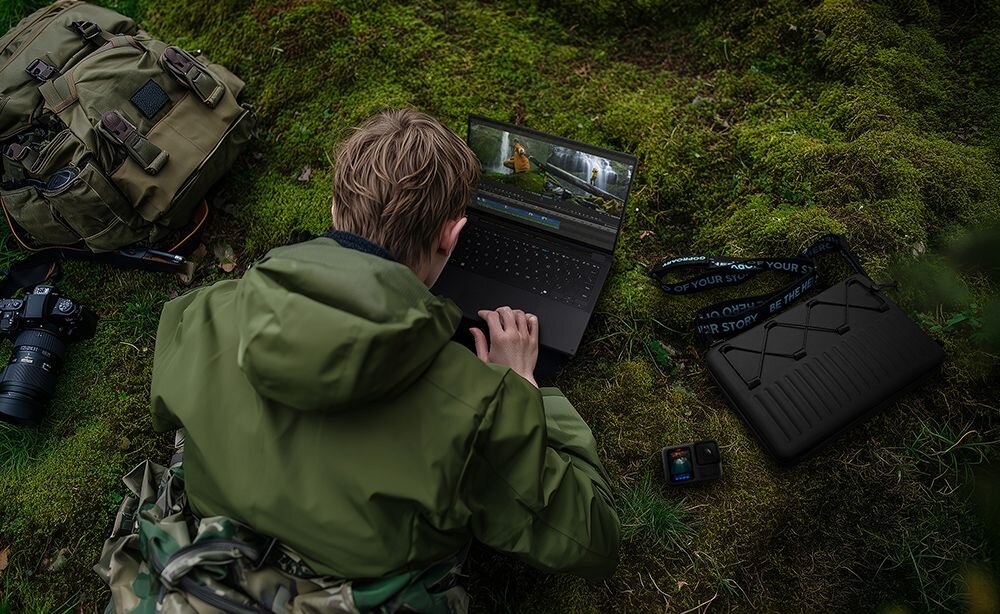 Osoba w zielonej kurtce pracuje na laptopie na tle zielonego mchu. Obok znajdują się plecak, aparat fotograficzny i etui na laptopa.