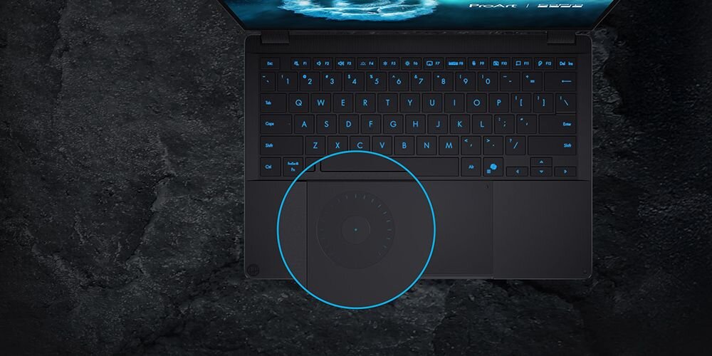Czarny laptop z podświetlaną na niebiesko klawiaturą i okrągłym panelem dotykowym. Na ekranie widoczna jest grafika z napisem \'ProArt\'.