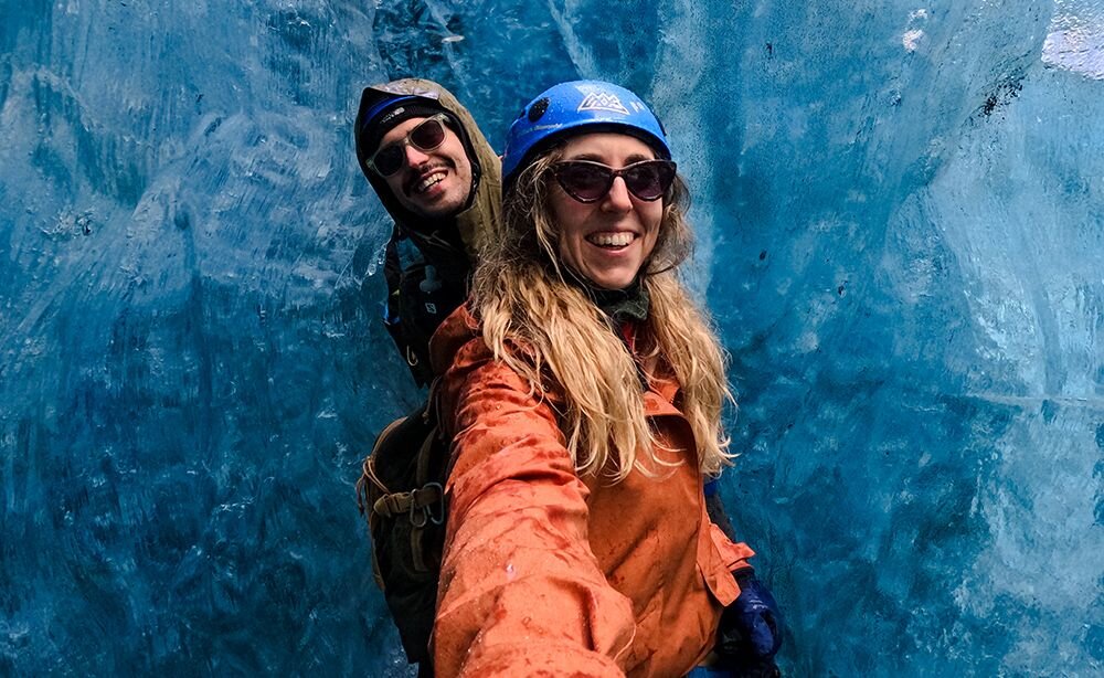 Dwie osoby w kaskach i okularach przeciwsłonecznych robią selfie na tle lodowej ściany. Osoby są ubrane w kurtki przeciwdeszczowe.