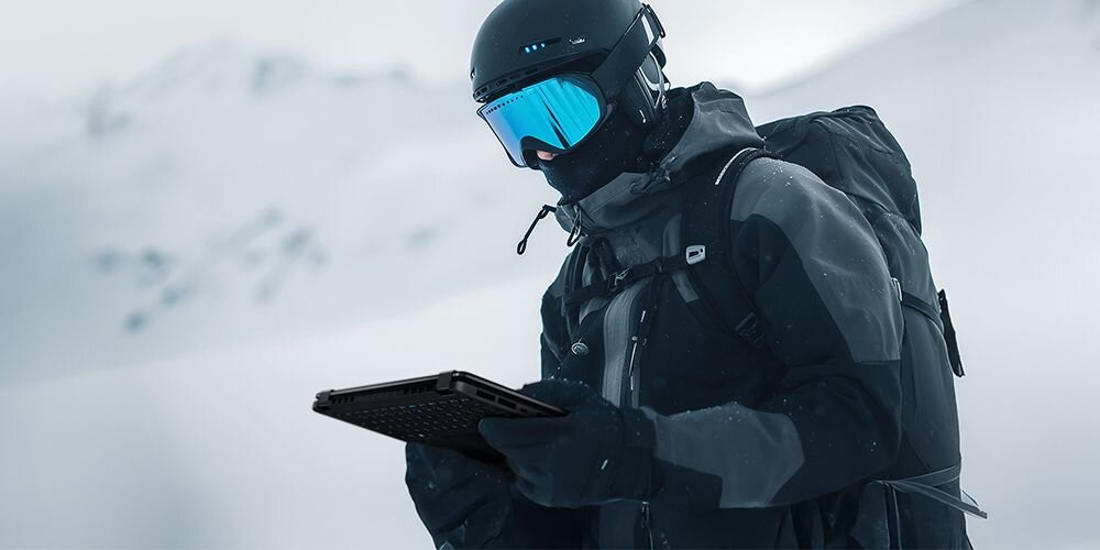 Osoba ubrana w zimową odzież ochronną trzyma laptopa na tle zaśnieżonego krajobrazu. Laptop jest czarny i ma widoczną klawiaturę.