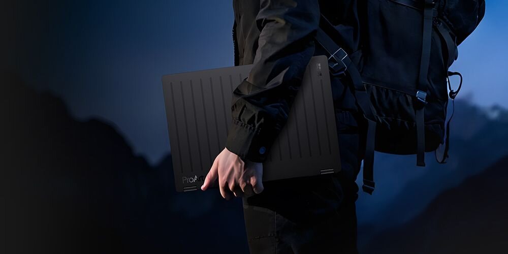 Osoba trzyma czarny laptop z widocznym napisem \'ProArt\' na tle górskiego krajobrazu o zmierzchu. Laptop ma pionowe linie na obudowie.