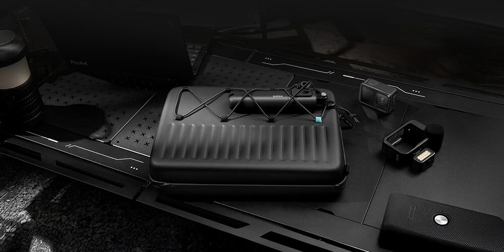 Na ciemnym biurku znajduje się czarny, prążkowany futerał z elastycznym paskiem, obok którego leży kamera sportowa i głośnik przenośny. W tle widoczny jest laptop z logo ProArt.