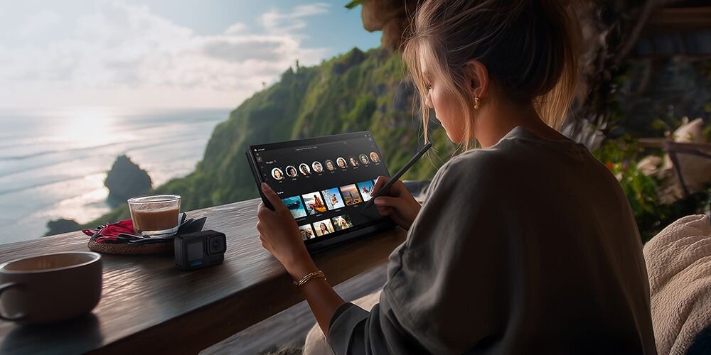 Osoba siedzi przy drewnianym stole, używając tabletu z rysikiem. W tle widać malowniczy widok na ocean i klify.