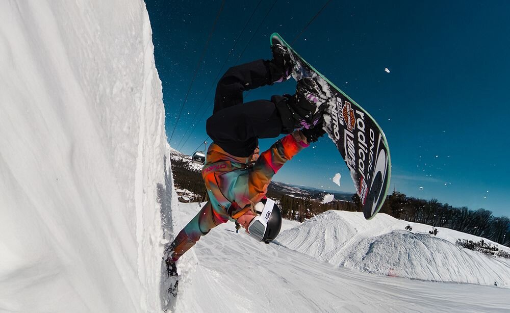 Osoba wykonuje akrobację na snowboardzie na śnieżnym stoku, ubrana w kolorową kurtkę i kask. W tle widać niebo i zaśnieżone wzgórza.