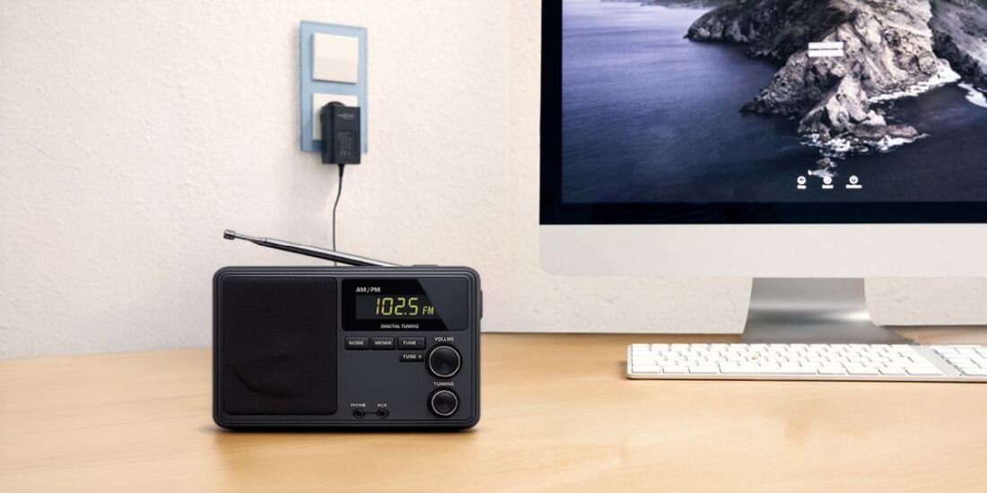 Na biurku znajduje się radio z wyświetlaczem pokazującym \'102.5 FM\'. W tle widać monitor komputera oraz zasilacz podłączony do gniazdka na ścianie.