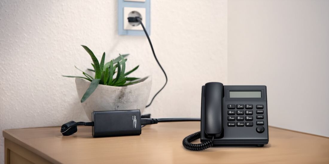 Na drewnianym blacie stoi czarny telefon stacjonarny z wyświetlaczem i klawiaturą numeryczną. Obok znajduje się zasilacz z widocznym logo \'ANSMANN\', a w tle doniczka z rośliną i gniazdko elektryczne.