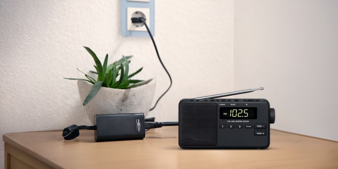 Na drewnianym blacie stoi czarne radio z wyświetlaczem pokazującym \'102.5\' oraz zasilacz podłączony do gniazdka. Obok znajduje się doniczka z zieloną rośliną.