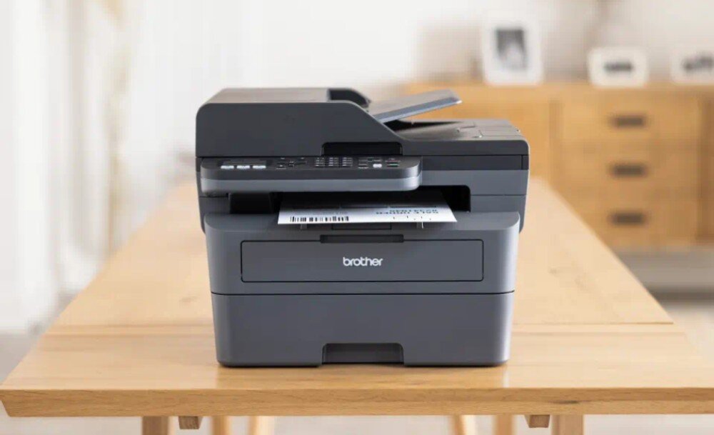 Urządzenie wielofunkcyjne BROTHER MFC-L2802DW urządzenie stojące przodem na stole z widoczną kartką wychodzącą z niego wygląd opis