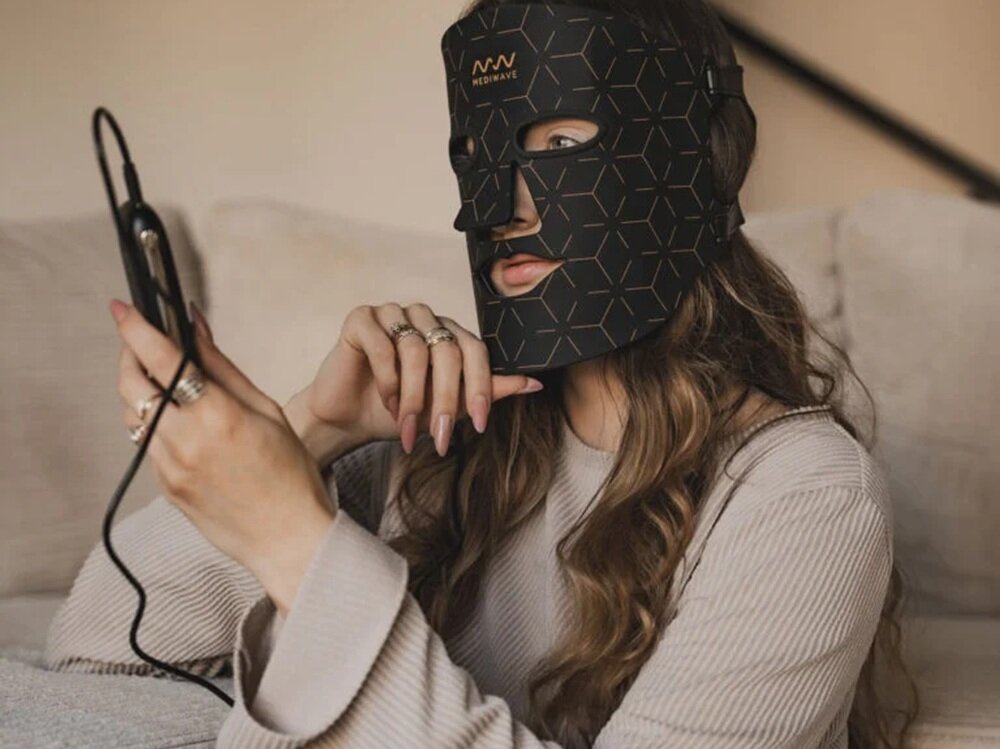 Osoba siedzi na kanapie, nosząc czarną maskę LED z geometrycznym wzorem i napisem \'MEDIWAVE\'. Trzyma w rękach urządzenie z kablem, patrząc na jego ekran.