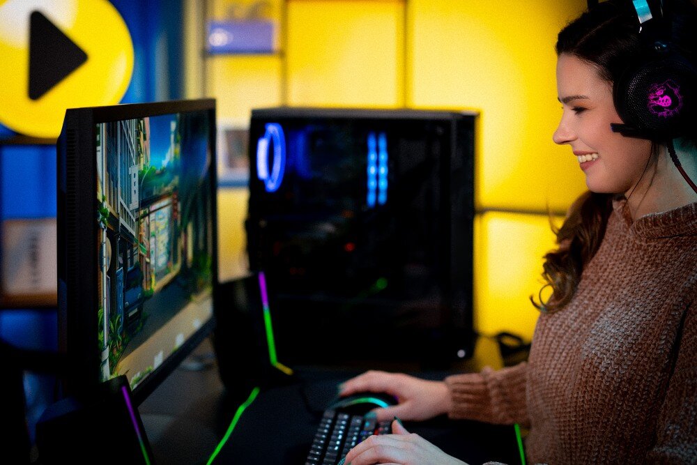 Komputer MAD DOG CORSAIR3500X-A02WR32WP Uśmiechnięta kobieta w słuchawkach korzystająca z komputera, siedząca przy biurku z widoczną w tle obudową z włączonym podświetleniem