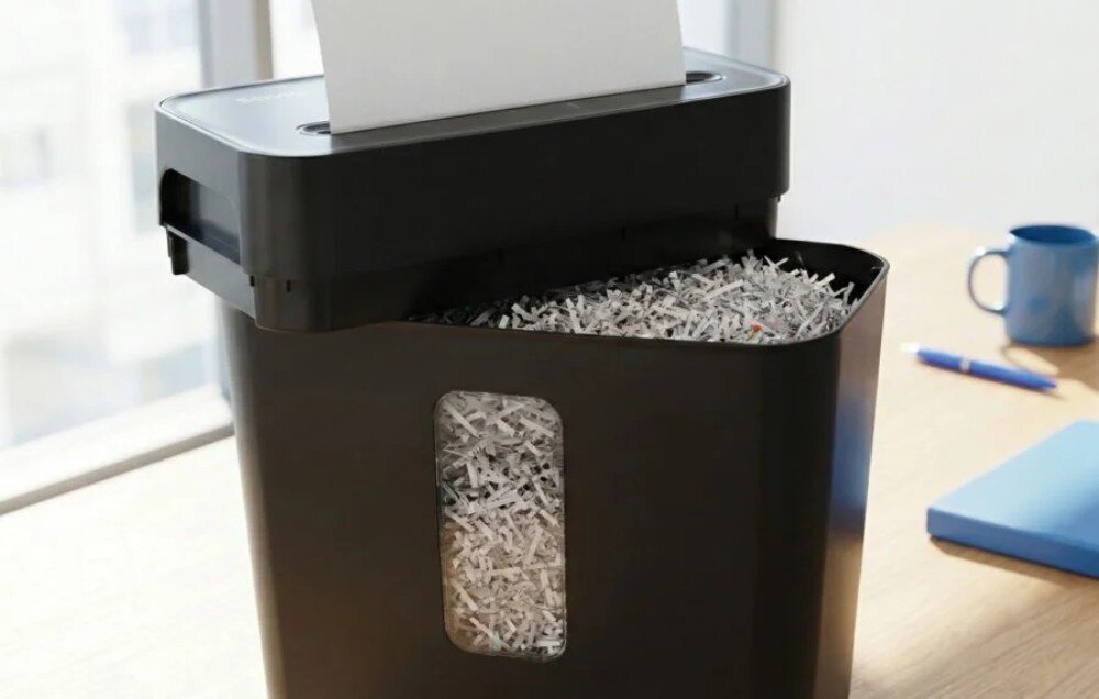 Niszczarka ścinkowa do dokumentów SAVIO ND-01 Zbliżenie na czarną niszczarkę z podniesioną górną częścią, pod którą widoczny jest kosz wypełniony jasnymi ścinkami papieru