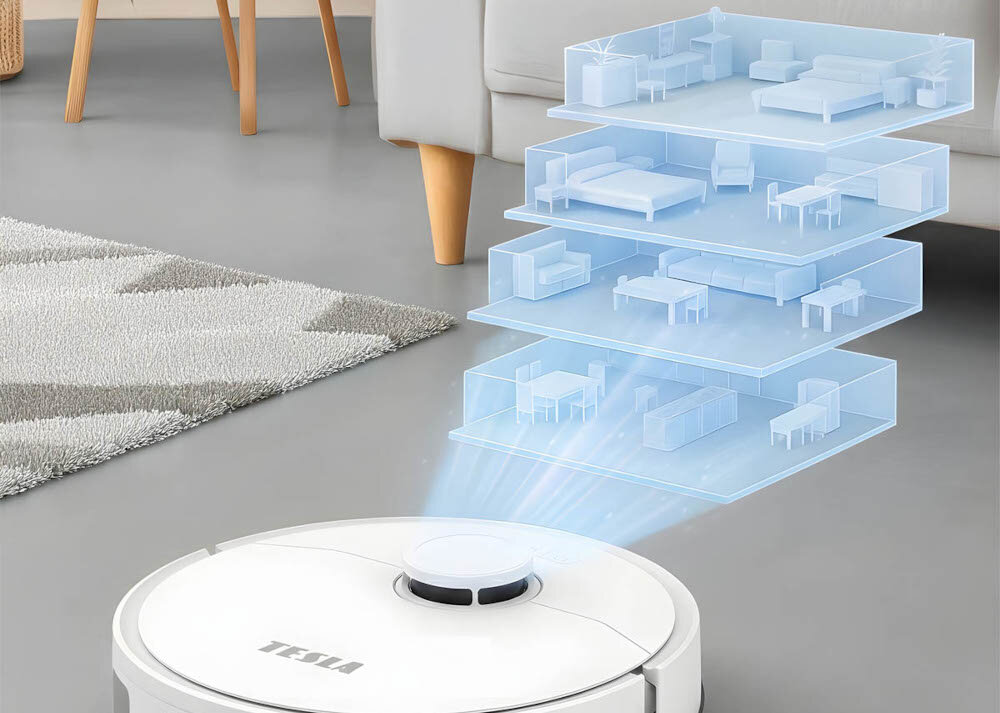 Na obrazie widoczny jest biały robot sprzątający z napisem \'TESLA\', z którego unosi się holograficzny model piętrowego mieszkania. W tle znajduje się fragment dywanu i noga kanapy.