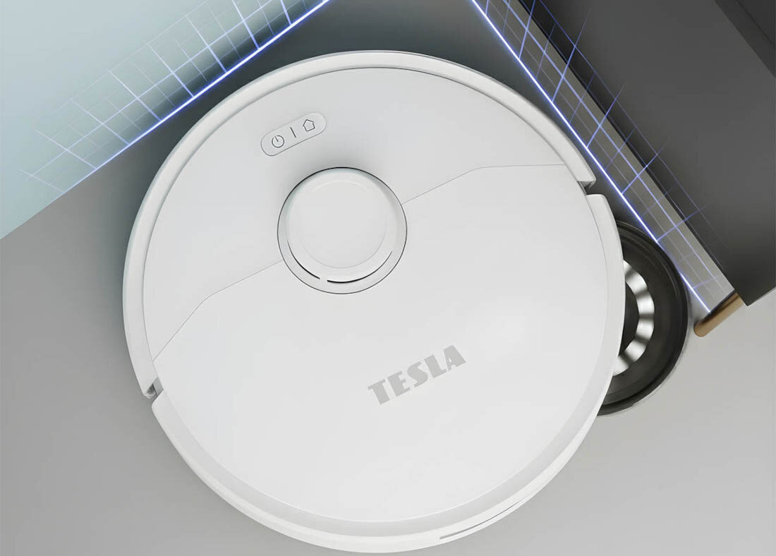 Na obrazie widoczny jest biały robot sprzątający z napisem \'TESLA\' na górze. Robot znajduje się w pobliżu narożnika pomieszczenia, a na jego powierzchni widoczne są dwa przyciski.